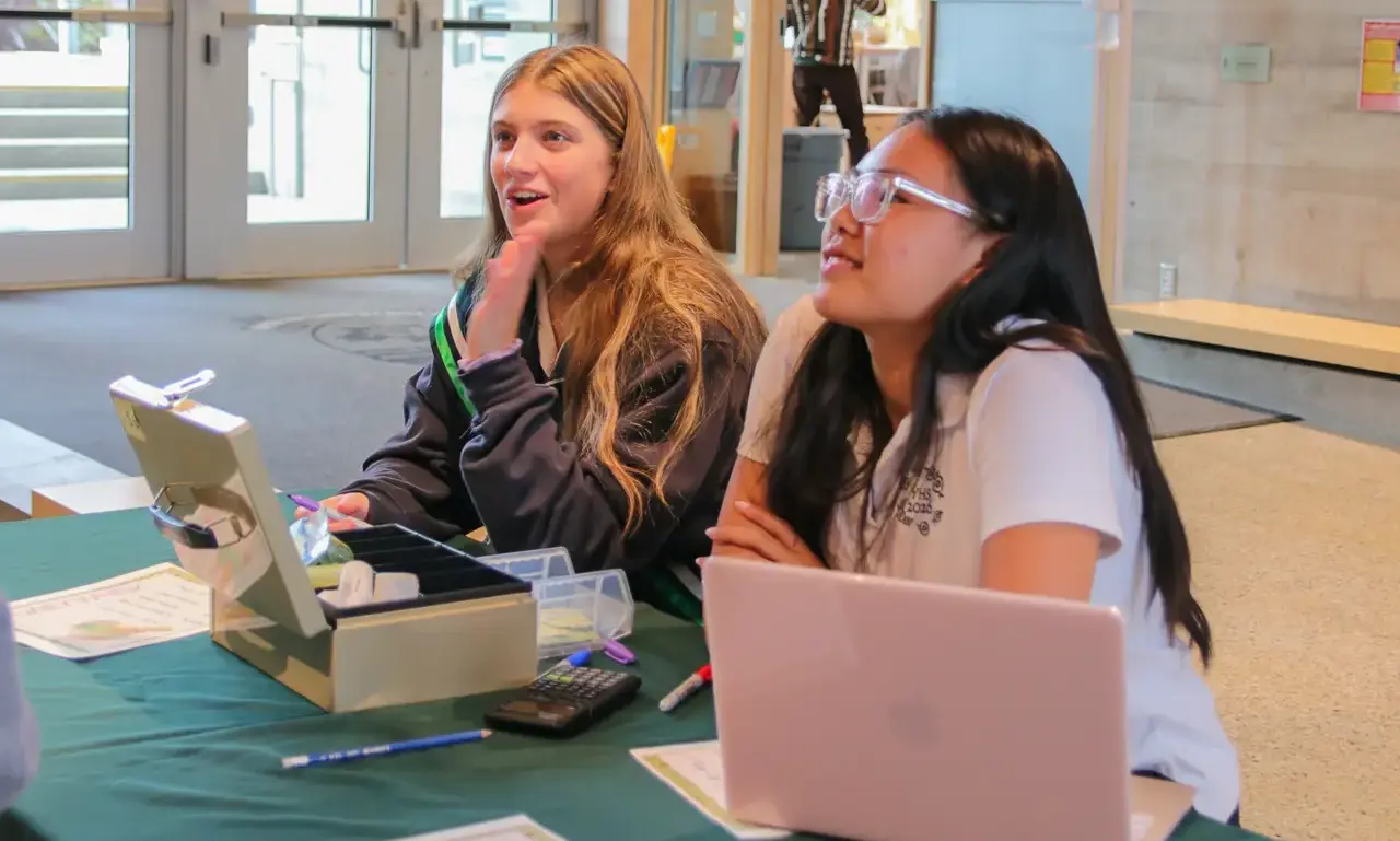 two secondary students sitting at a table behind a laptop and a cash box